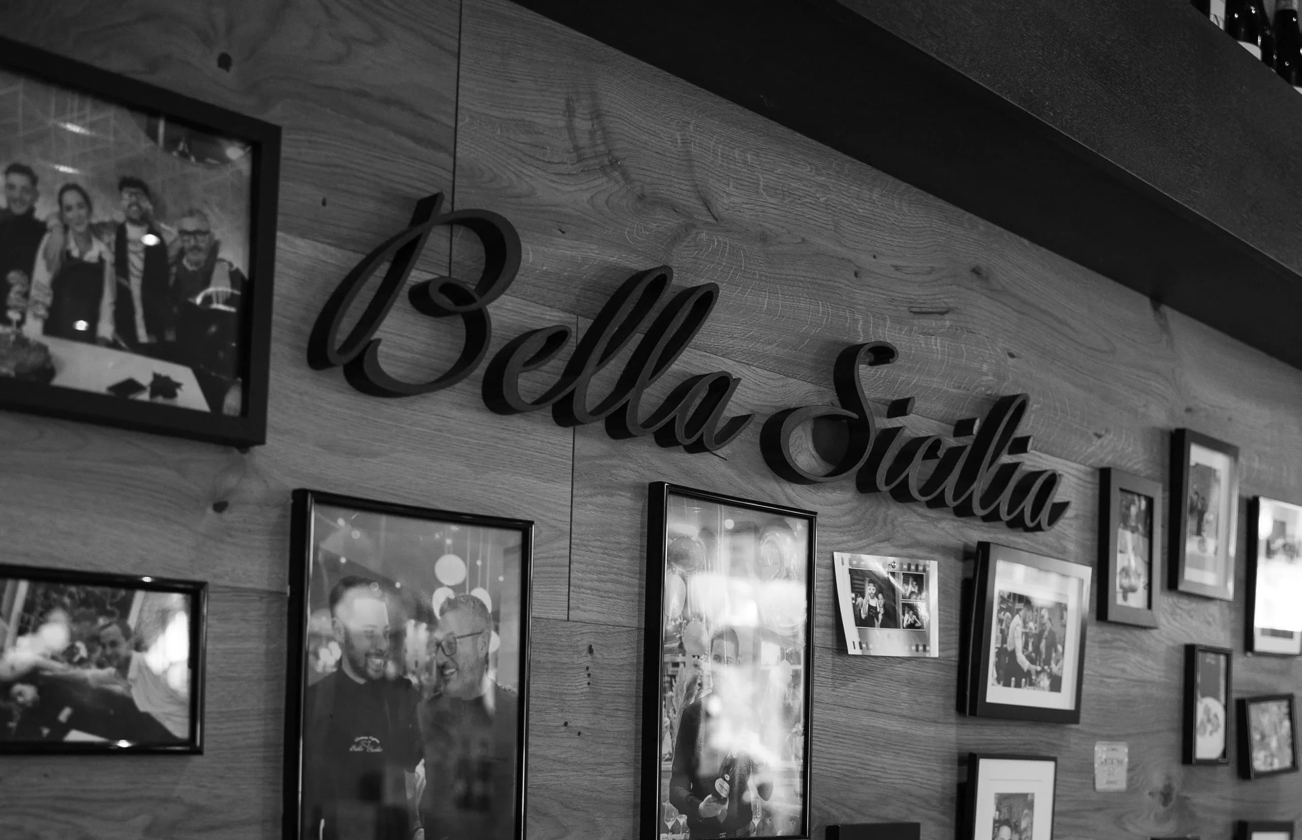 Die Wand im Gastraum mit dem Schriftzug Bella Sicilia und gerahmten Fotos, schwarzweiß