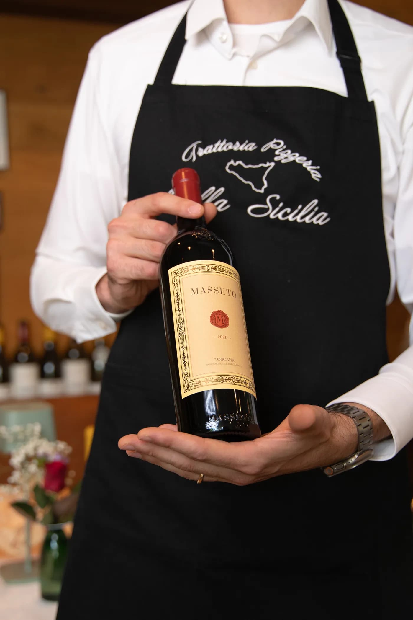 Eine Flasche Masseto, vor der Schürze mit dem Schriftzug Bella Sicilia gehalten